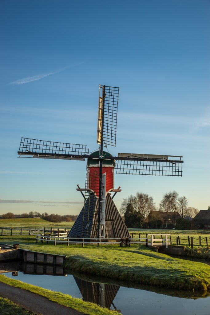 A vertical shot of a post mill on a grassy field with wooden fences and a blue sky in the background